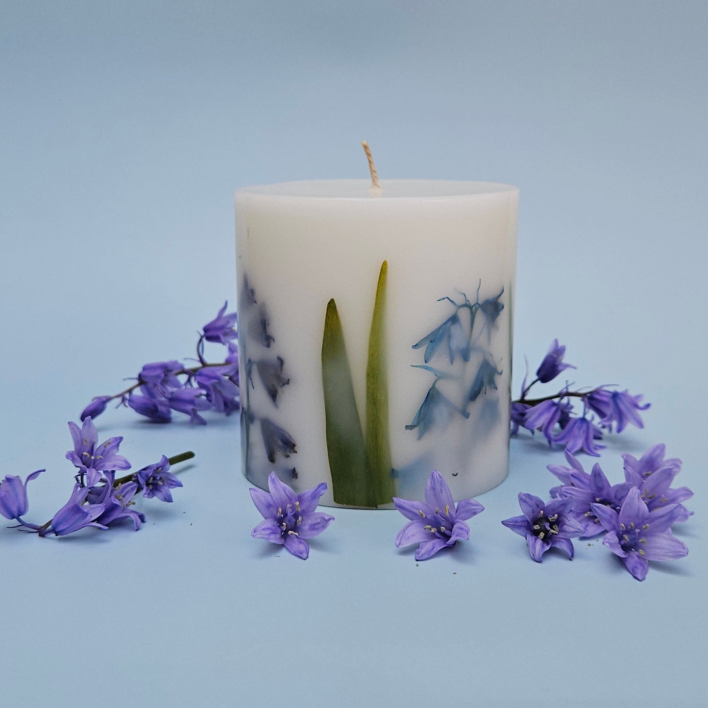 A botanical candle featuring real bluebells and leaves embedded in the wax. The candle is encircled by bluebell flowers against a soft, light blue background.