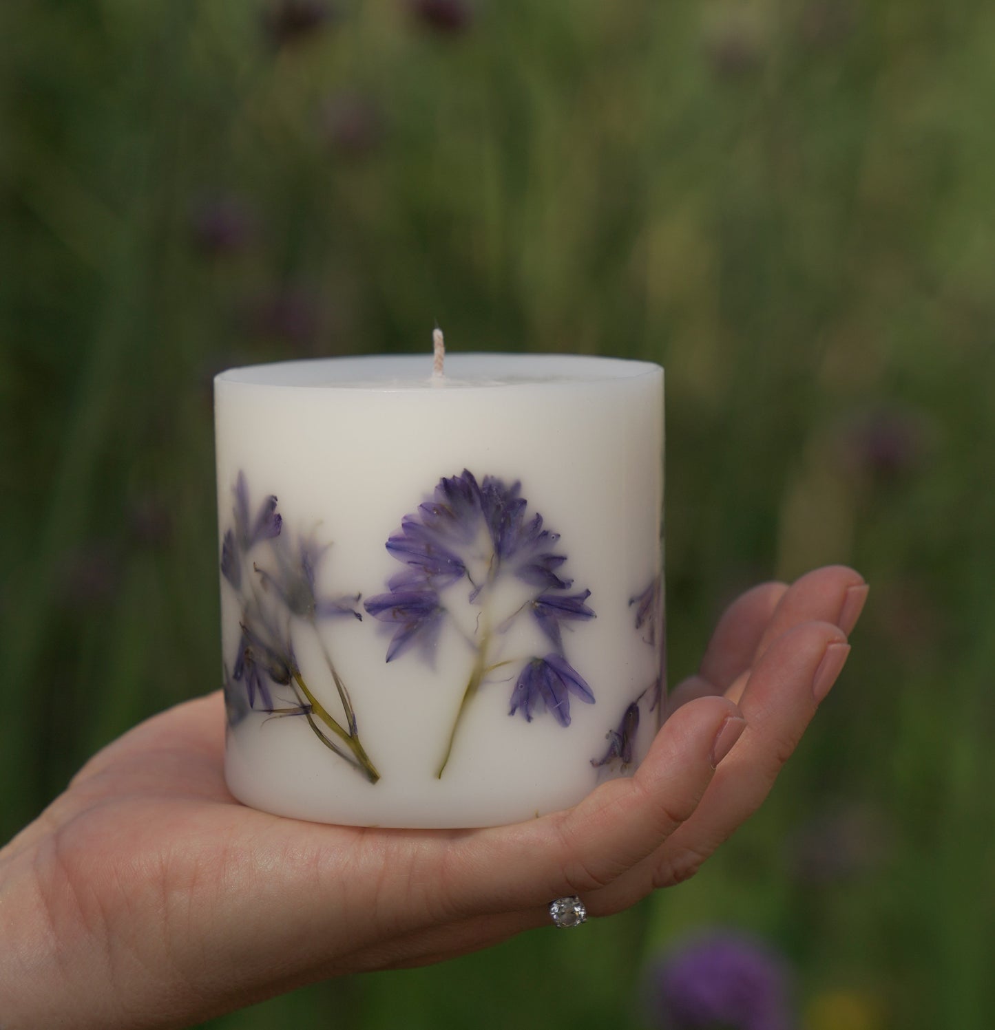 A bluebell botanical candle with real bluebells on its sides, held in a hand with a ring on the finger.