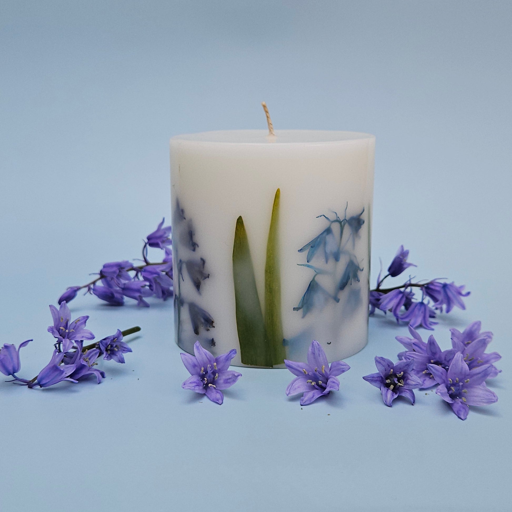 A botanical candle featuring real bluebells and leaves embedded in the wax. The candle is encircled by bluebell flowers against a soft, light blue background.