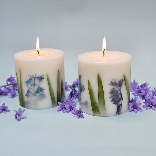 A pair of lit botanical candles with real bluebell flowers embedded within the wax, surrounded by loose bluebell blooms on a light blue background.