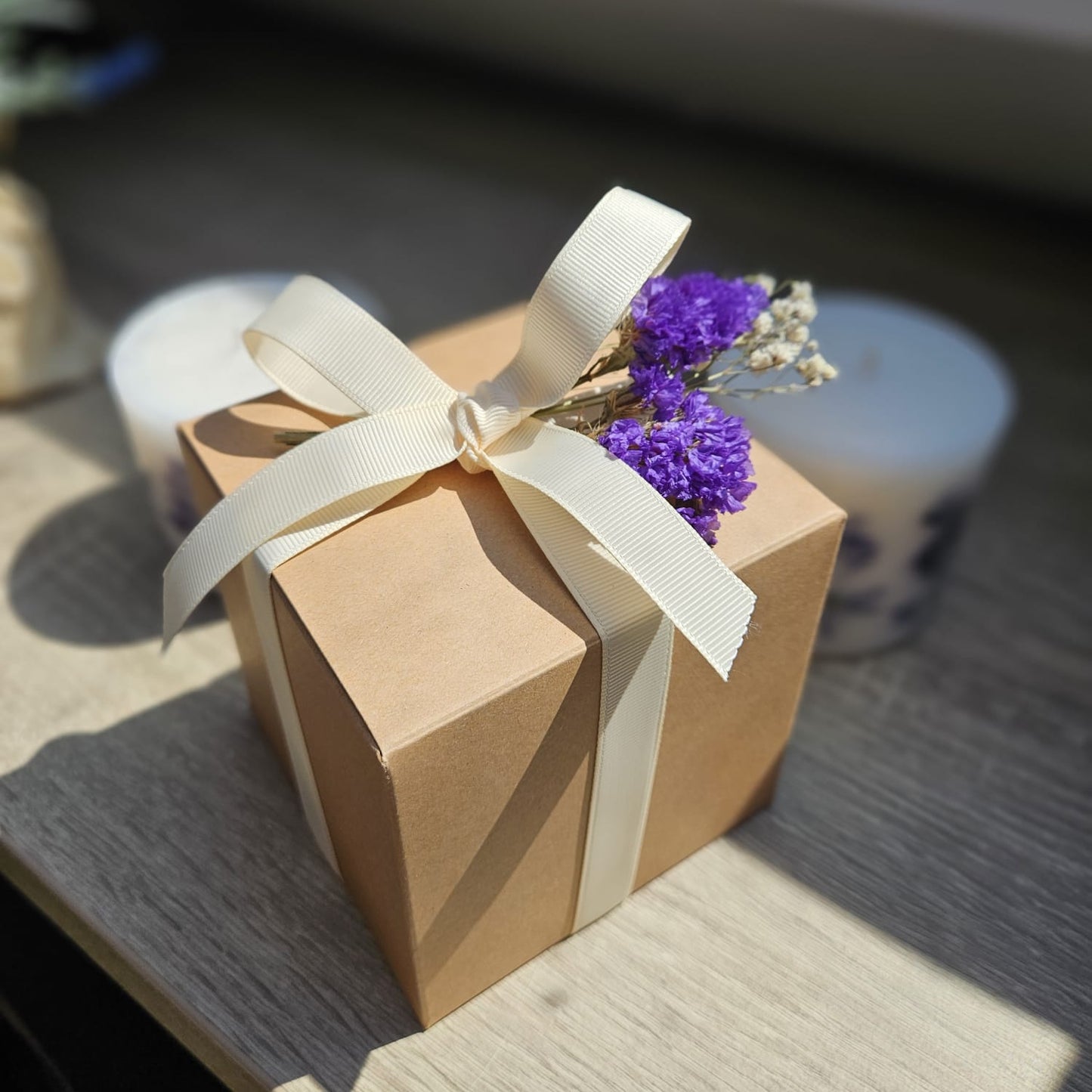 A kraft paper box with a bow and dried flowers, standing on a wooden shelf with light falling on it.
